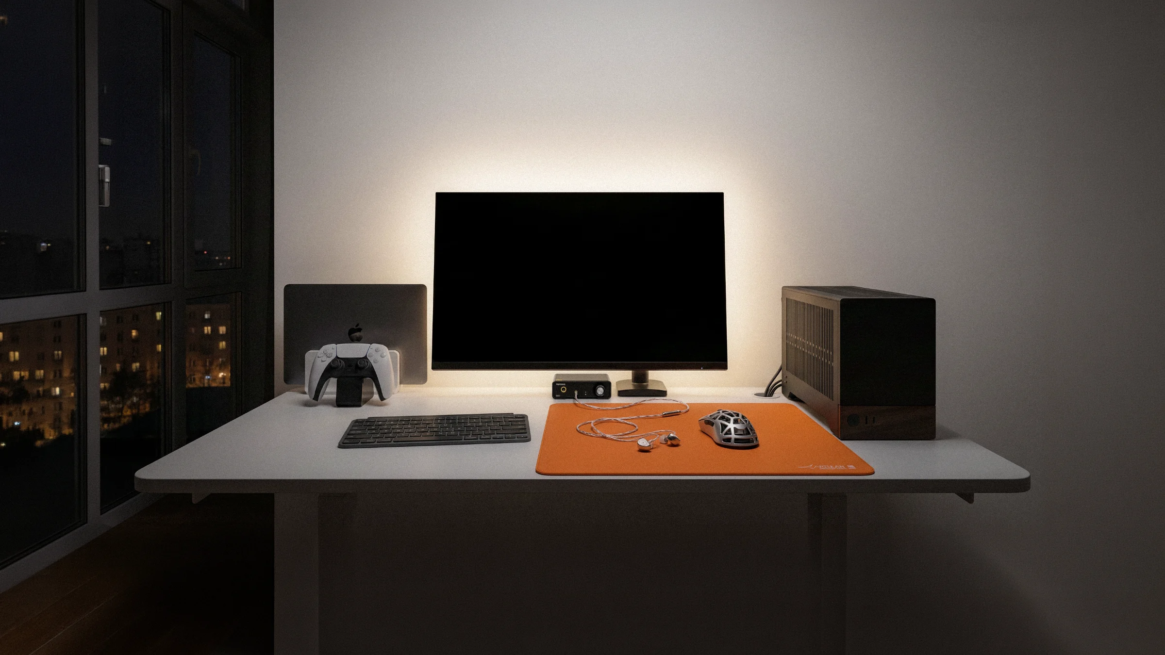 A dark room, a lit desk: monitor, keyboard, orange mat, a small PC and a laptop.